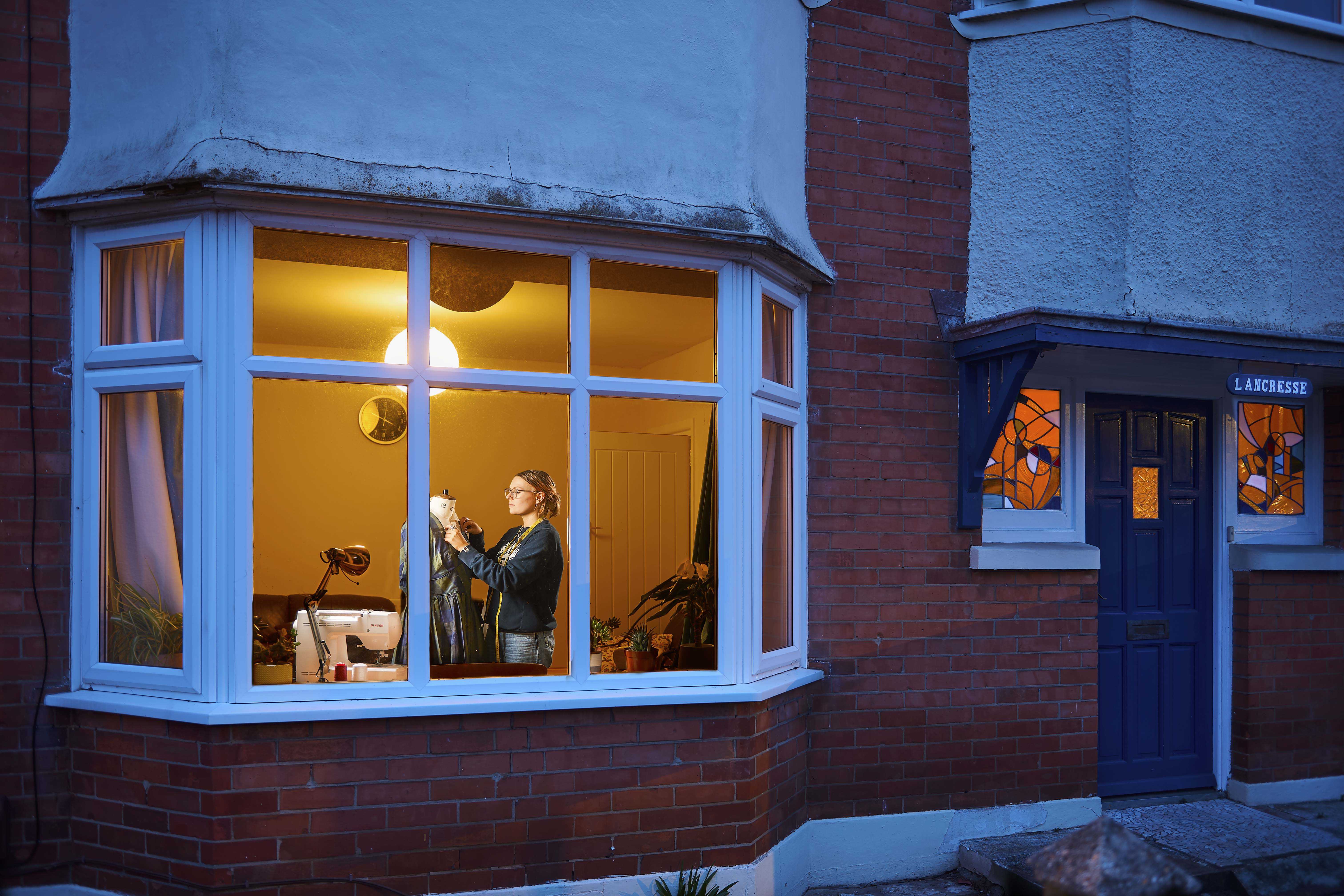 A woman photographed from outside a house; she is dressing a mannequin with a sewing machine next to her.