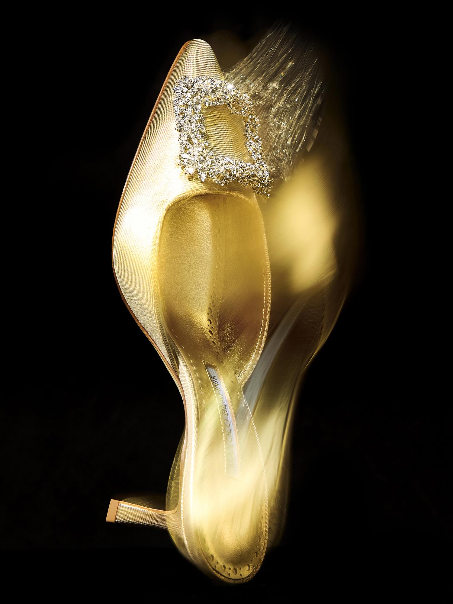 A gold satin high-heeled shoe with a decorative rhinestone buckle on its toe. The shoe is positioned against a black background, highlighting its shimmering surface and elegant design.