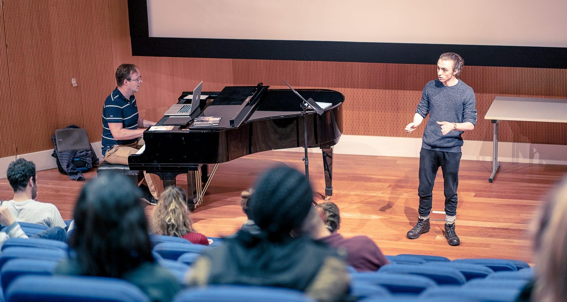 A person sits playing a piano while another person stands talking to an audience.