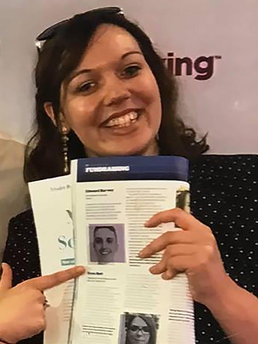 A smiling woman holds a magazine page to the camera and points at it.