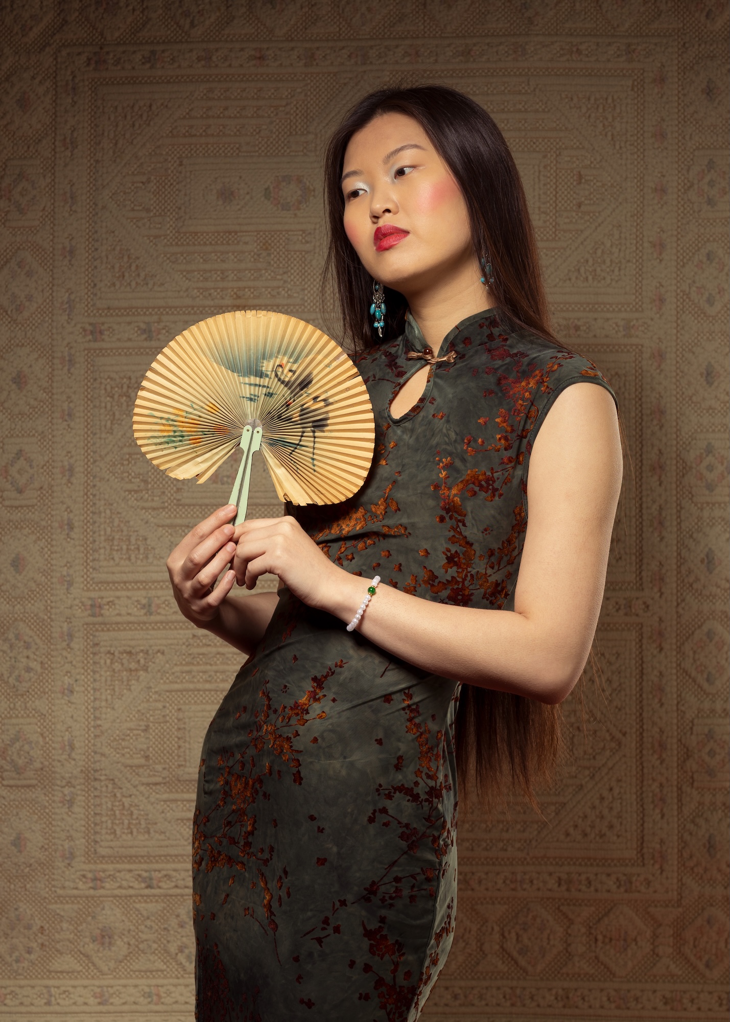 A fashion model wearing a long, elegant black and bronze dress and holding a small Japanese-style fan poses for a photo.