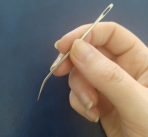 A hand holding a gold curved sewing needle against a dark blue background.