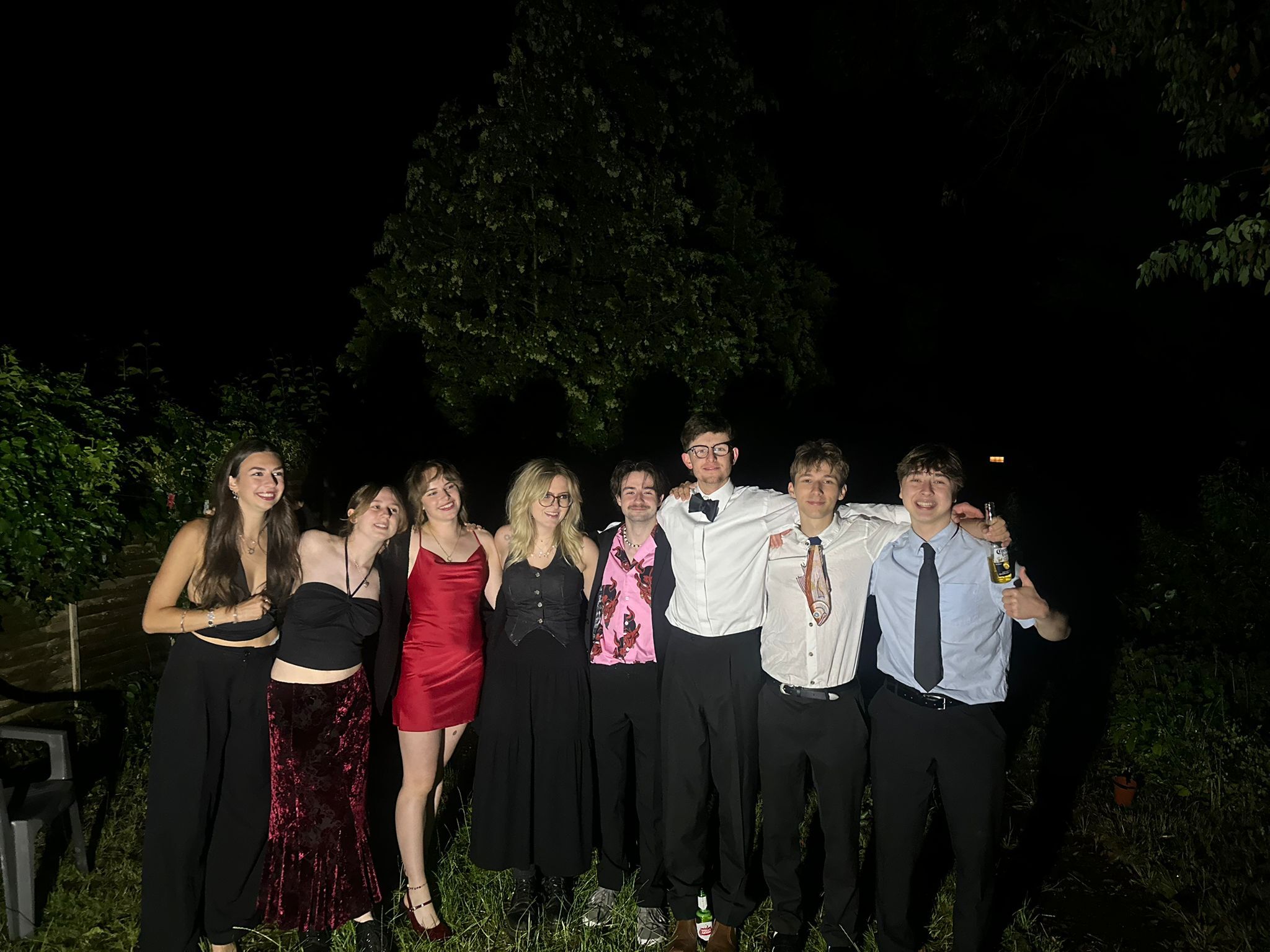 Group of nine people in formal attire posing outdoors at night, with dark trees and foliage in background.