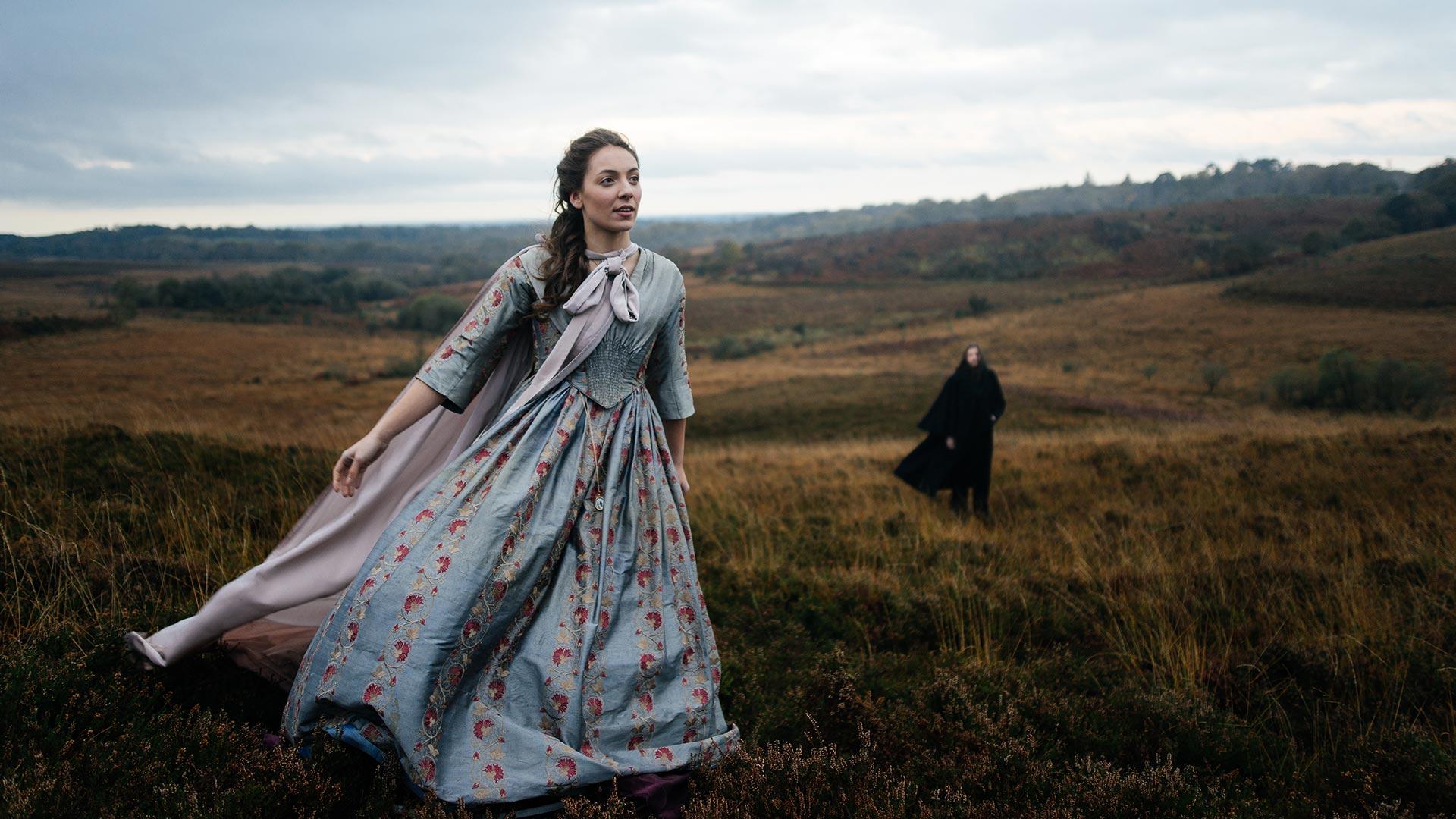 A woman in period costume walking across a grassy moor. A man, also in period costume, stands far in the background and watches.