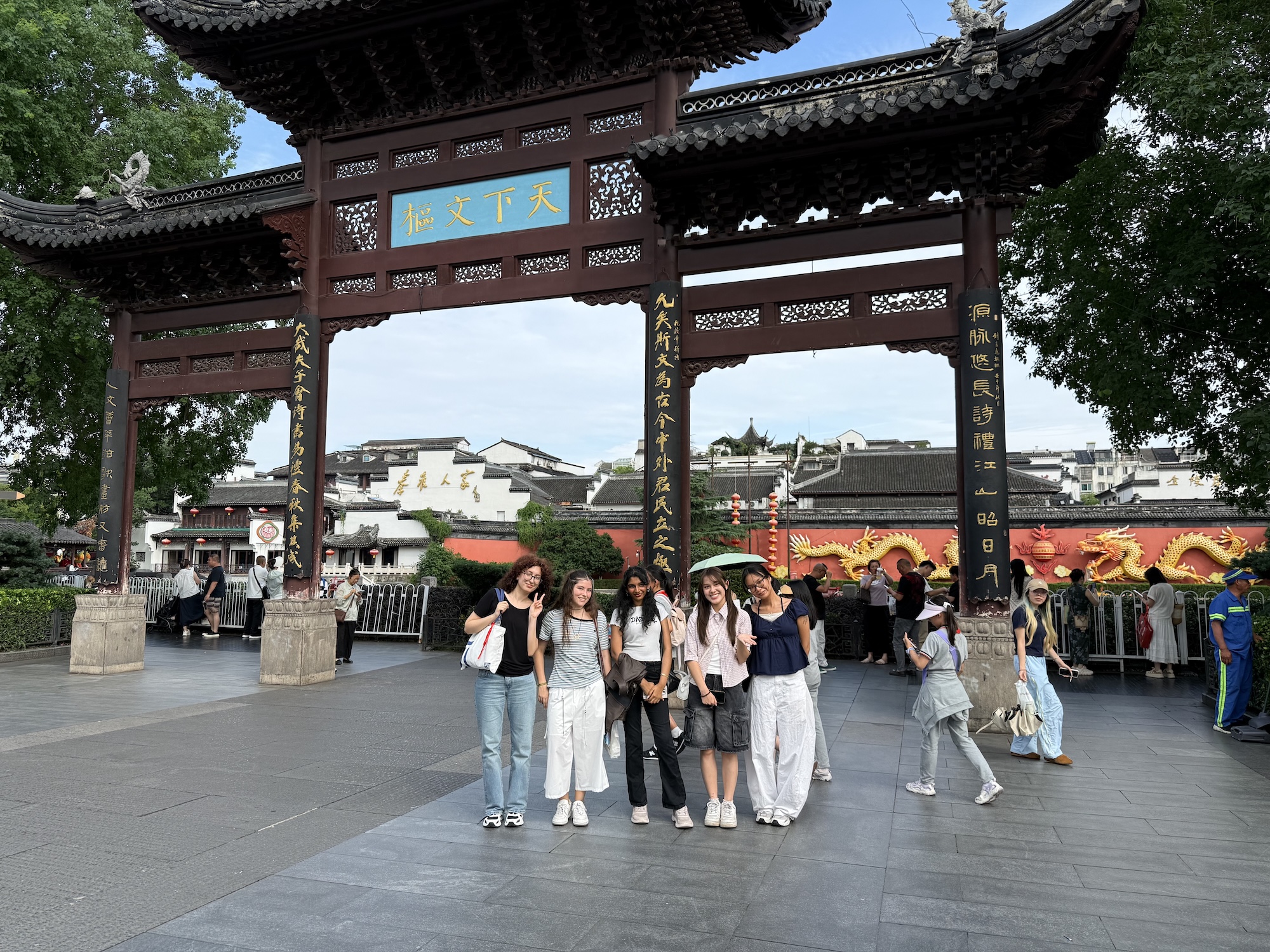 A group of six young women pose for a photo in front of a large traditional Chinese archway with red and black features. The background displays a bustling street scene with people and shops under a clear sky.