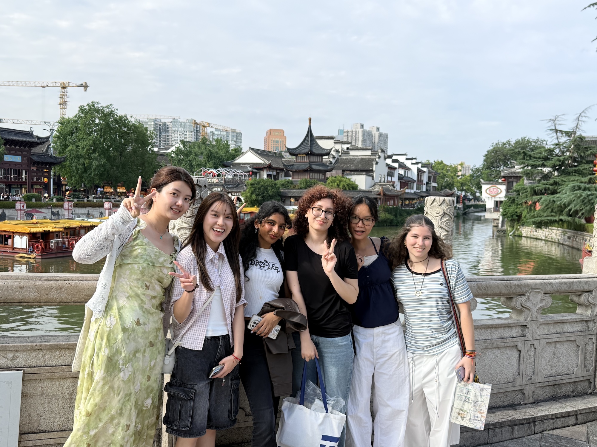 A group of six people poses by a river in a city setting. They stand on a stone railing, smiling and making peace signs. Behind them, traditional architectural features are visible along the water, with modern buildings in the distance.