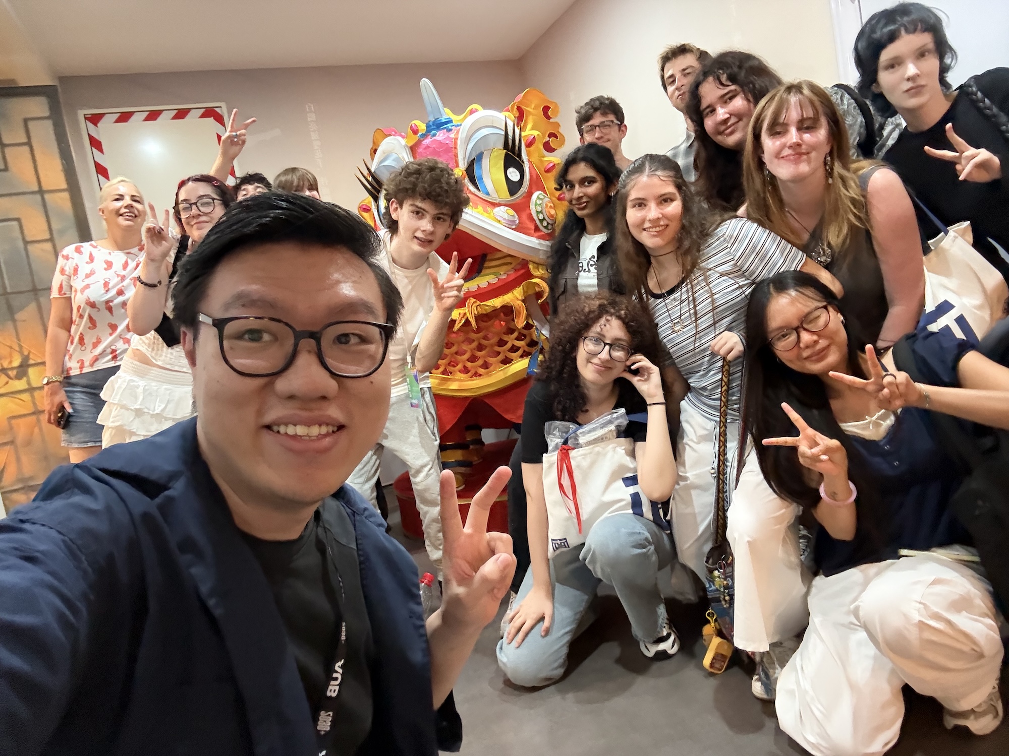 A group of young people posing together for a photograph in front of a large, colourful dragon statue. The scene is bright, with a minimalist background, and the individuals are smiling and making peace signs.