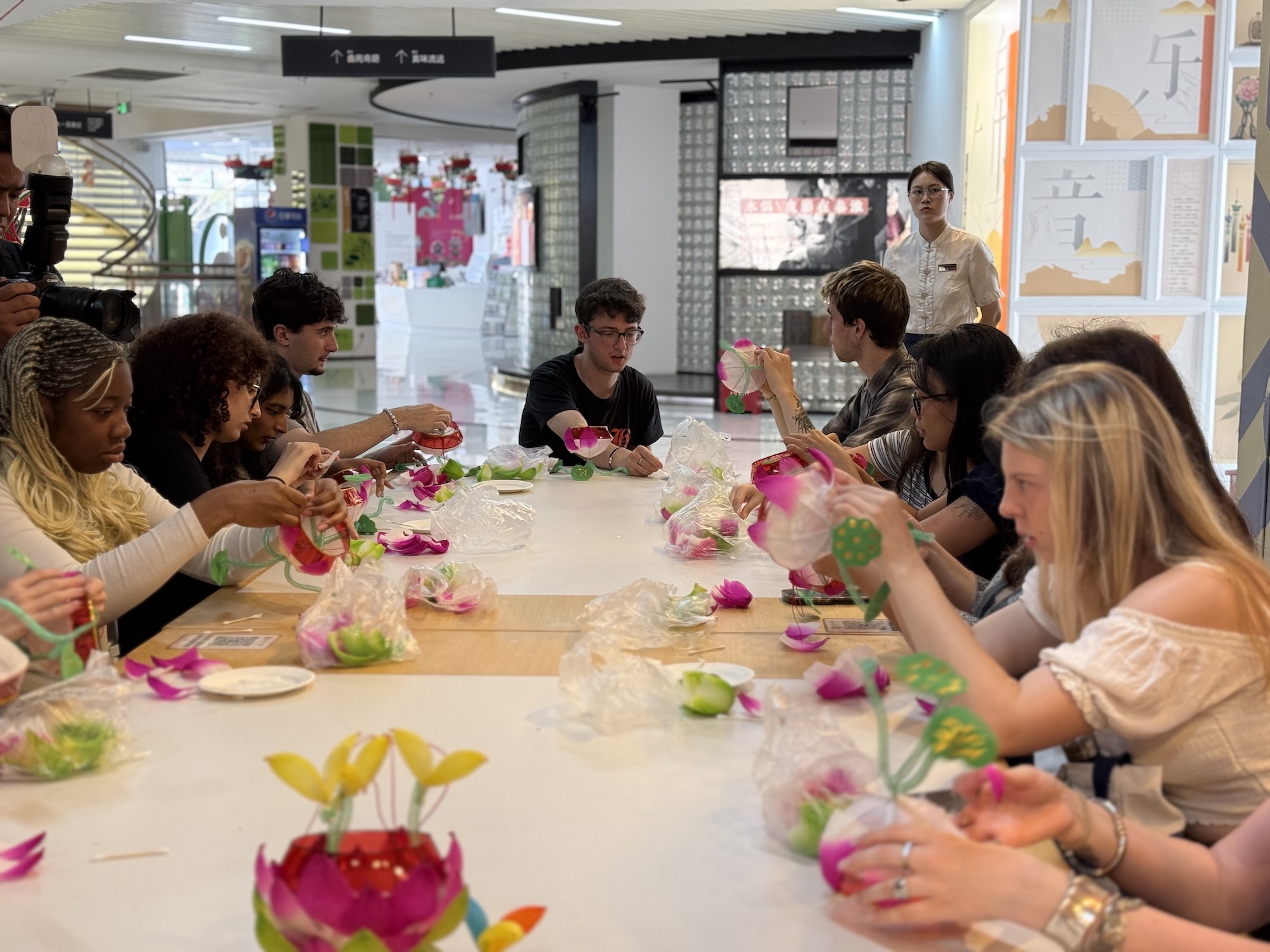 A group of young people seated around a long table engaged in a craft activity, creating flower arrangements. Various colourful materials, including pink petals and green leaves, are visible on the table. An instructor observes in the background.