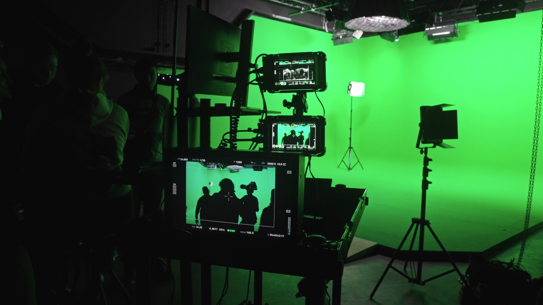Film studio with bright green screen backdrop, camera equipment on tripods, and monitors displaying footage in dark room.