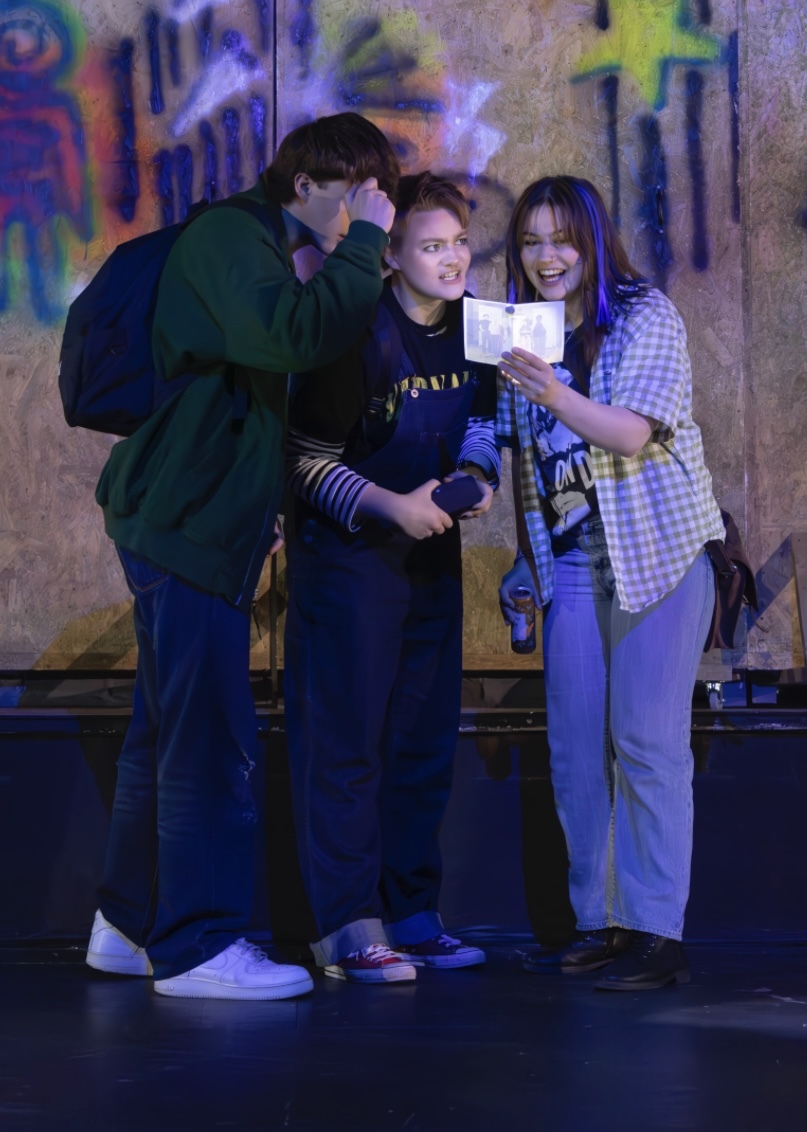 Three young people looking at mobile phone together against colourful graffiti wall, wearing casual clothing in evening light.