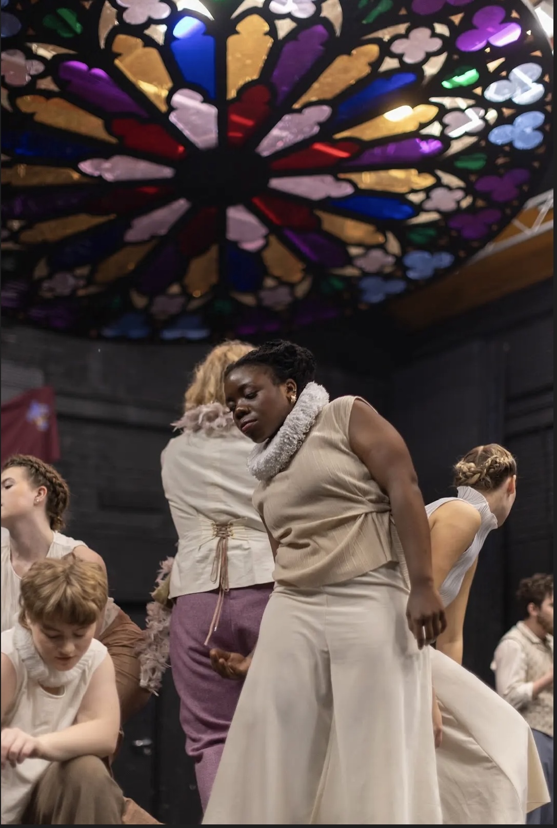 Two actors embrace centre stage beneath ornate circular stained glass ceiling featuring radiating multicoloured panels in blues, purples, oranges and pinks.