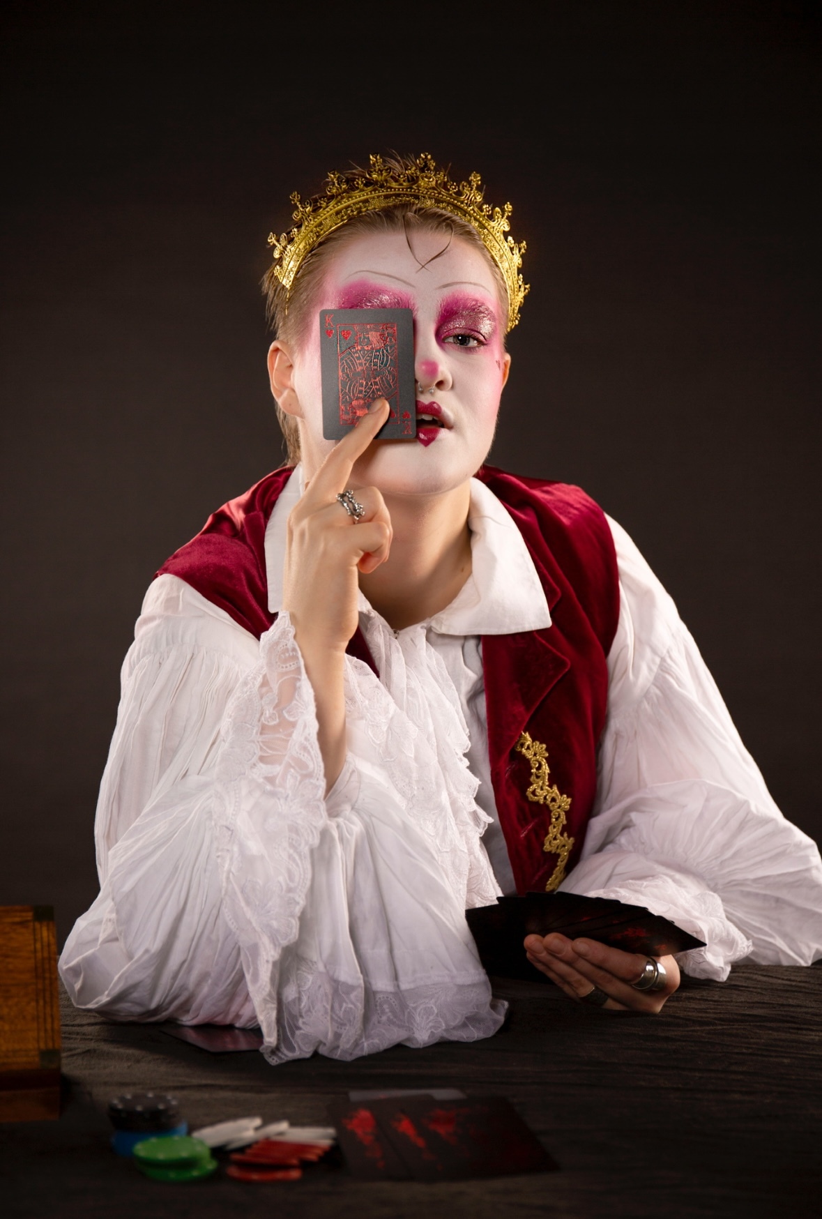 Person in theatrical makeup with gold crown, red and white costume, holding playing card to face against dark background.