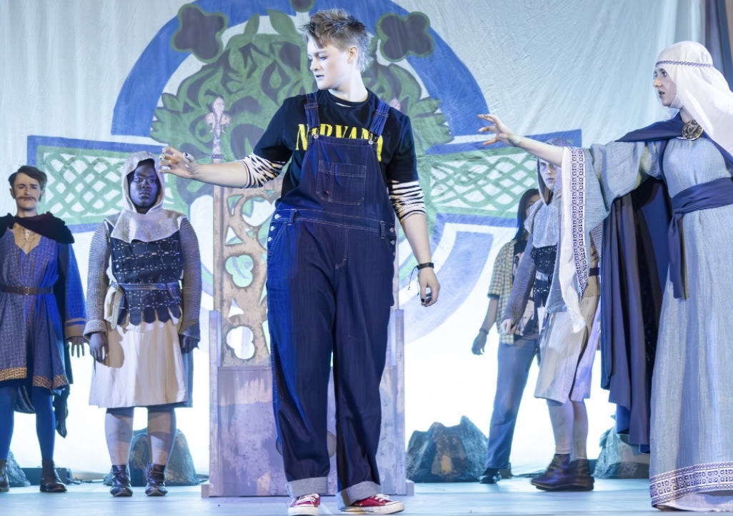 Young performer in black dungarees centre stage with cast members in medieval costumes on colourful set backdrop.