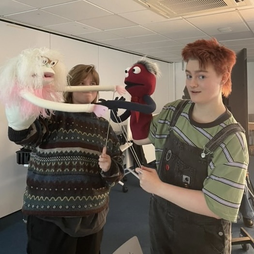 Two people operating puppet characters in white office space. Person on left wears patterned jumper, person on right wears striped shirt.