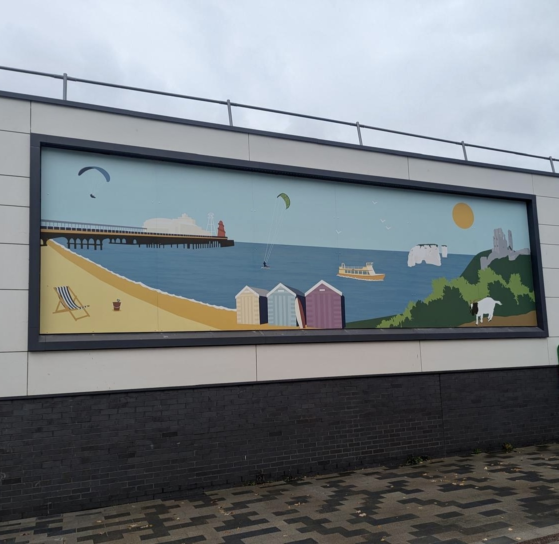 Large mural on grey building wall depicting blue sea, yellow sand, colourful beach huts, green hills, white sheep, and orange sun.