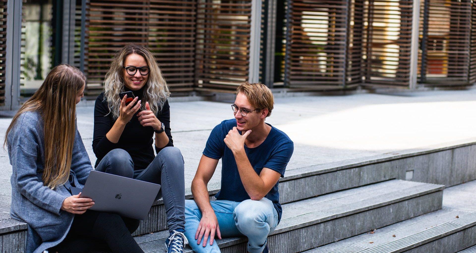 A group of three people sat on steps looking at a laptop and talking, the person in the middle is looking at a mobile phone