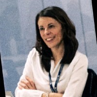 Woman with long dark hair in white blazer, arms crossed, smiling at camera against light blue background.