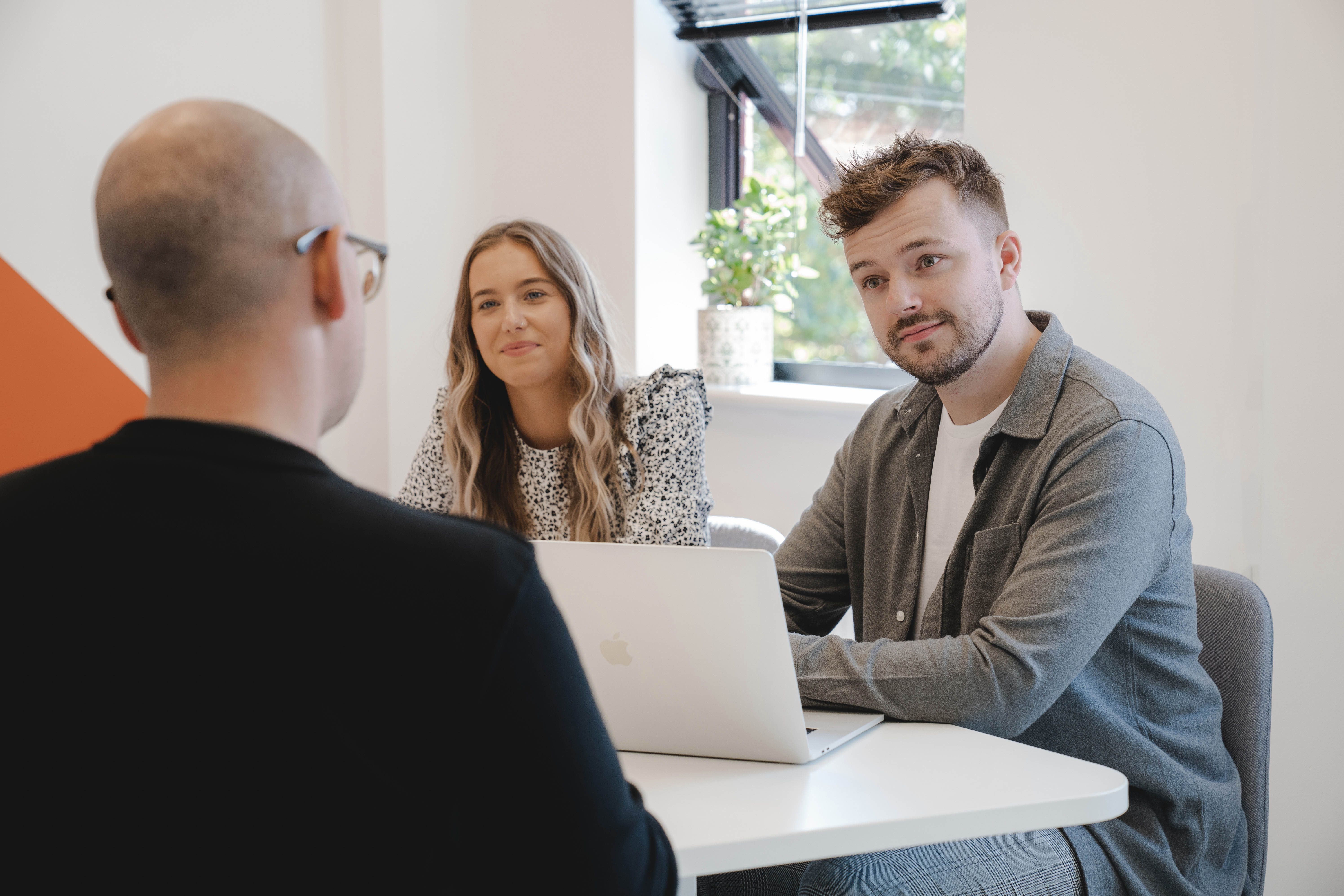 Two people sit behind a desk with a laptop open in front of them and speak with a visitor