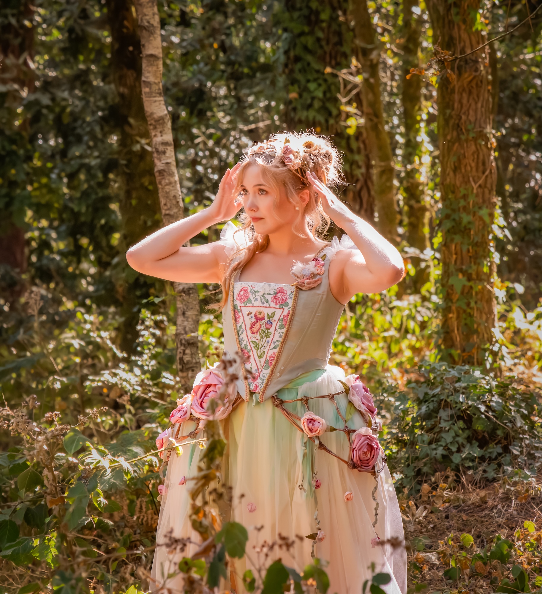 A woman in a floral, corset-style gown stands in a wooded setting with sunlit trees and greenery.