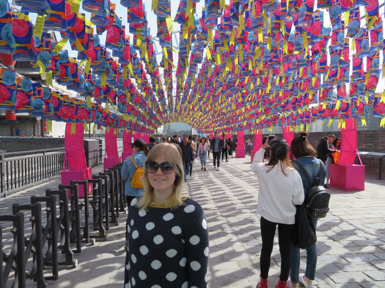 A blonde woman wearing sunglasses and a black polka-dot top stands on a paved walkway under rows of colourful hanging flags, with other pedestrians and railings in the background.