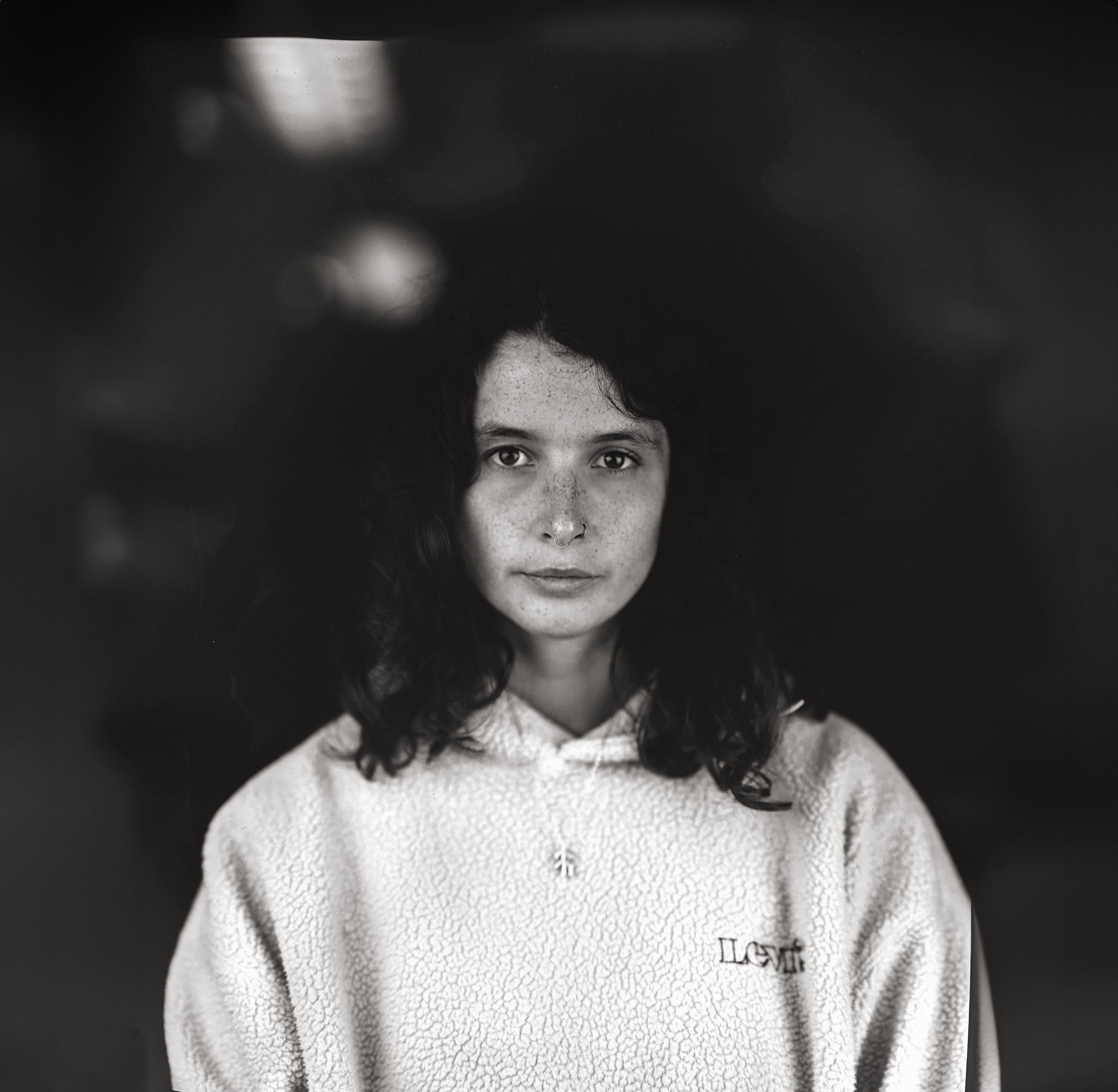 Black and white portrait of woman with dark wavy hair wearing light-coloured hoodie, looking directly at camera against dark background.