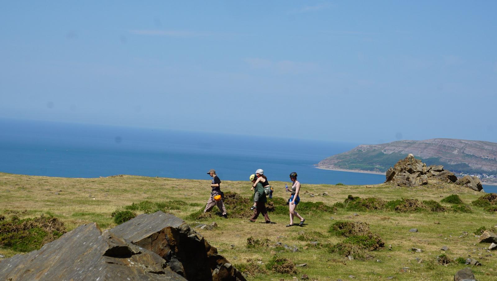 Four hikers walking across grassy clifftop with blue sea and distant headland visible under clear sky.