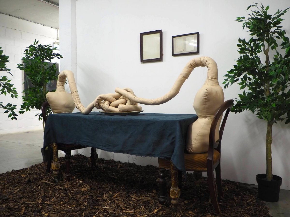 A sculpture of two objects sitting at a table connected by a long tube, twisted and knotted in the middle.