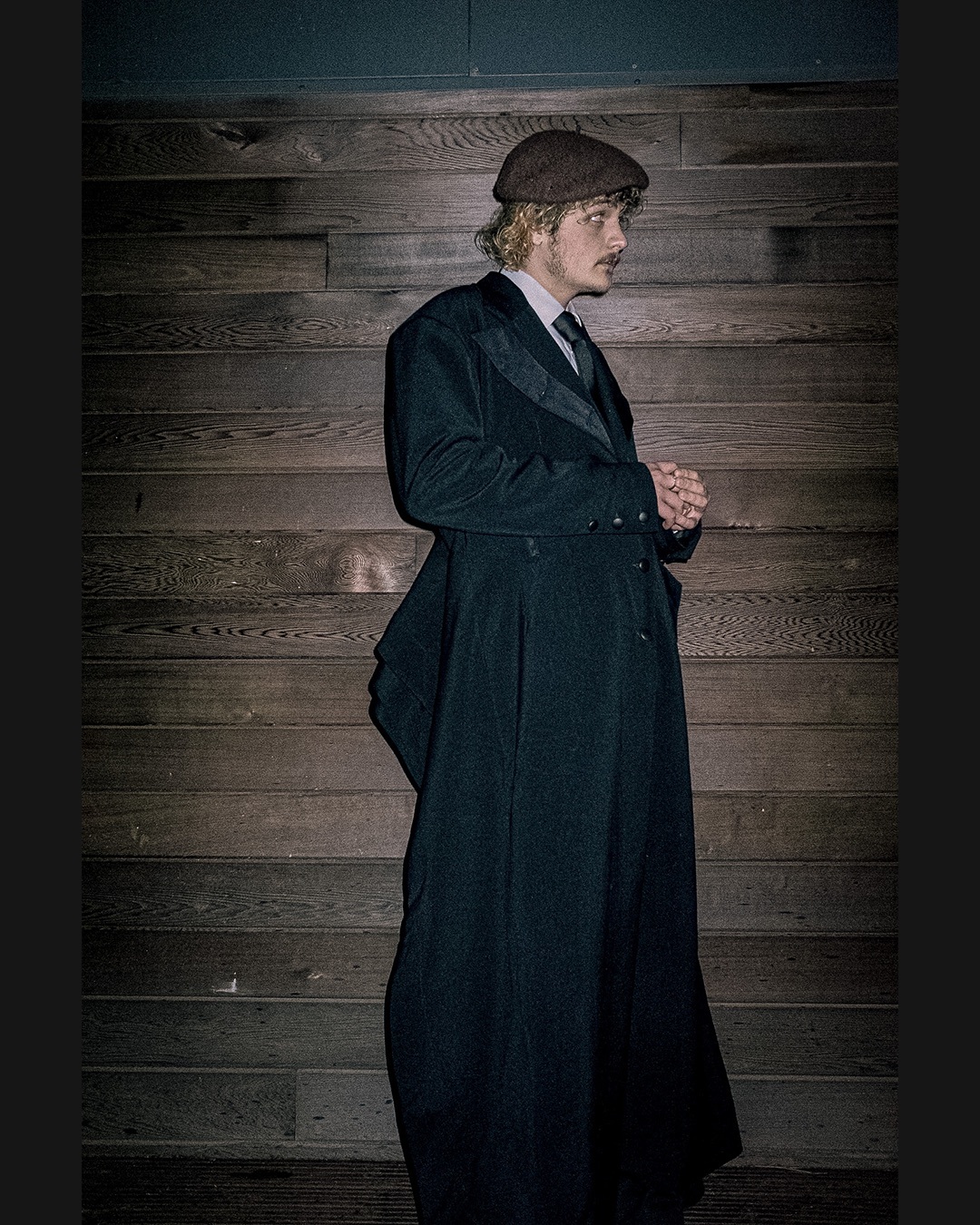 A man in a long black coat in front of a wood background