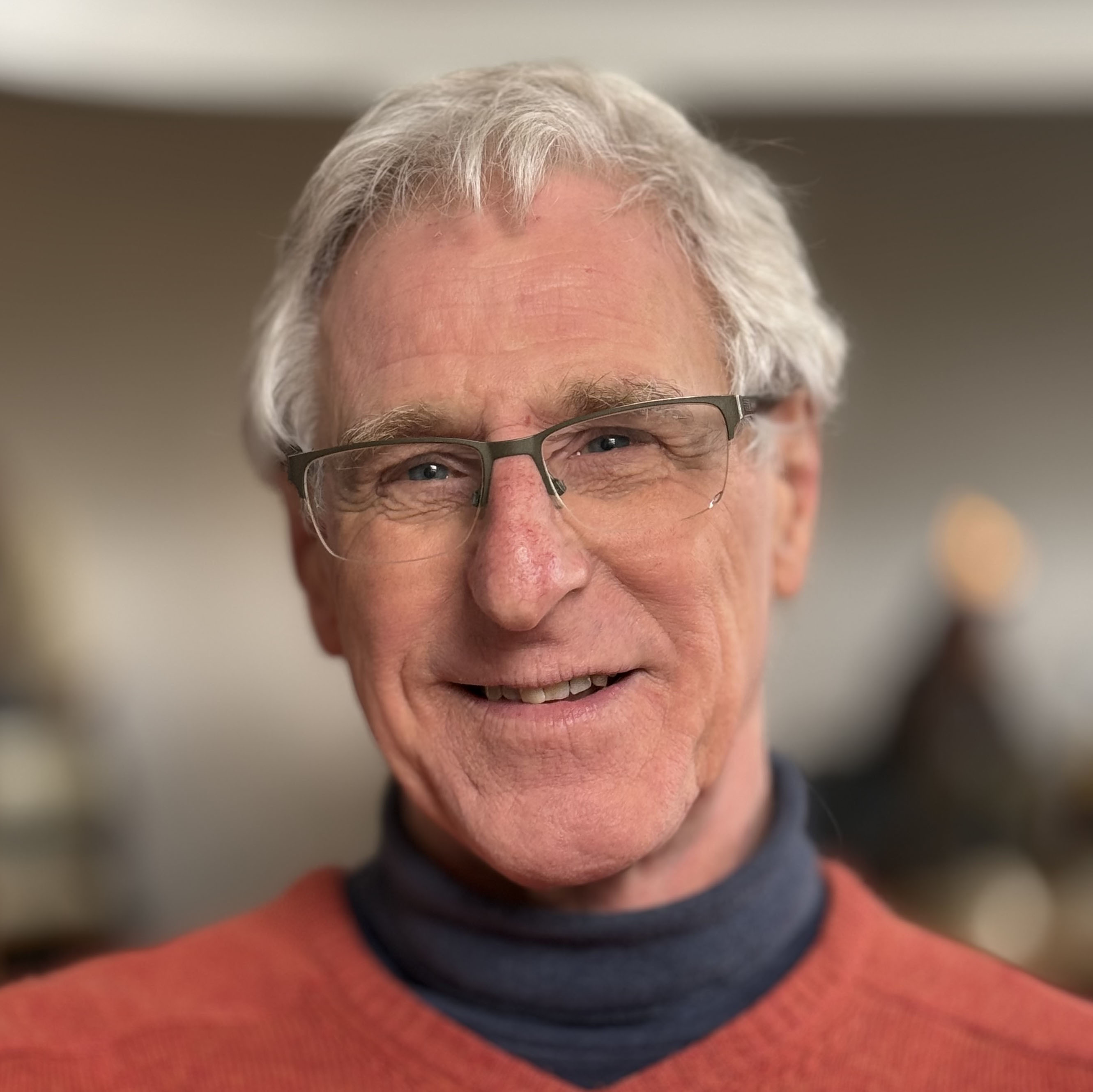 Elderly man with grey hair and glasses wearing red jumper over navy turtleneck, smiling at camera indoors
