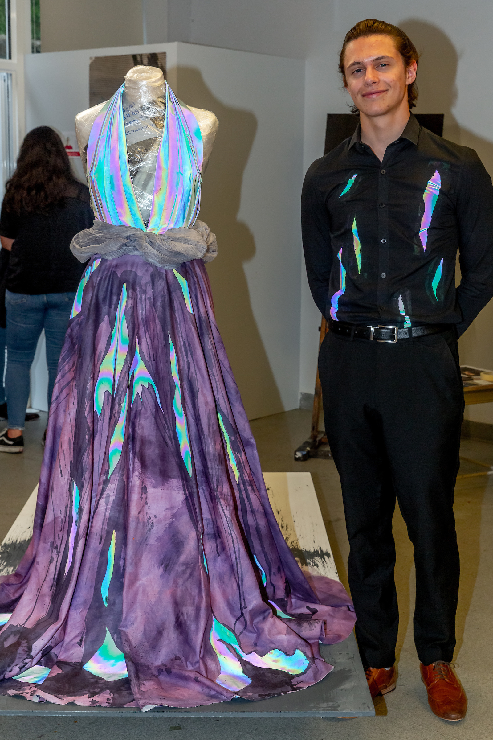 Alex Matheson stood next to the iridescent dress dress he designed in an exhibition