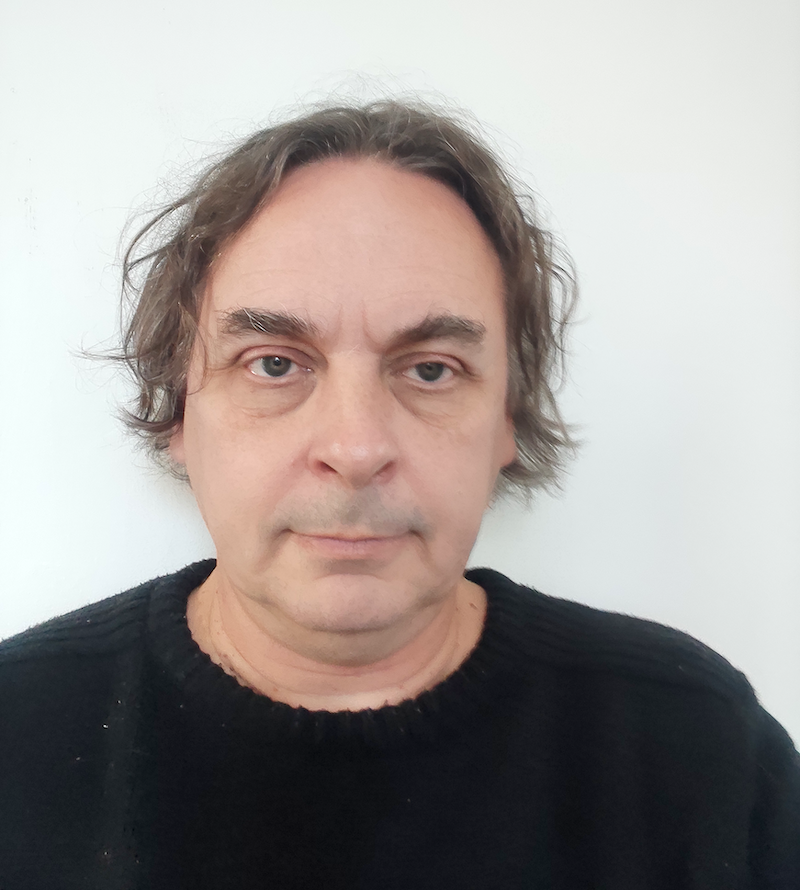 Middle-aged man with brown wavy hair and green eyes wearing black jumper against white background.
