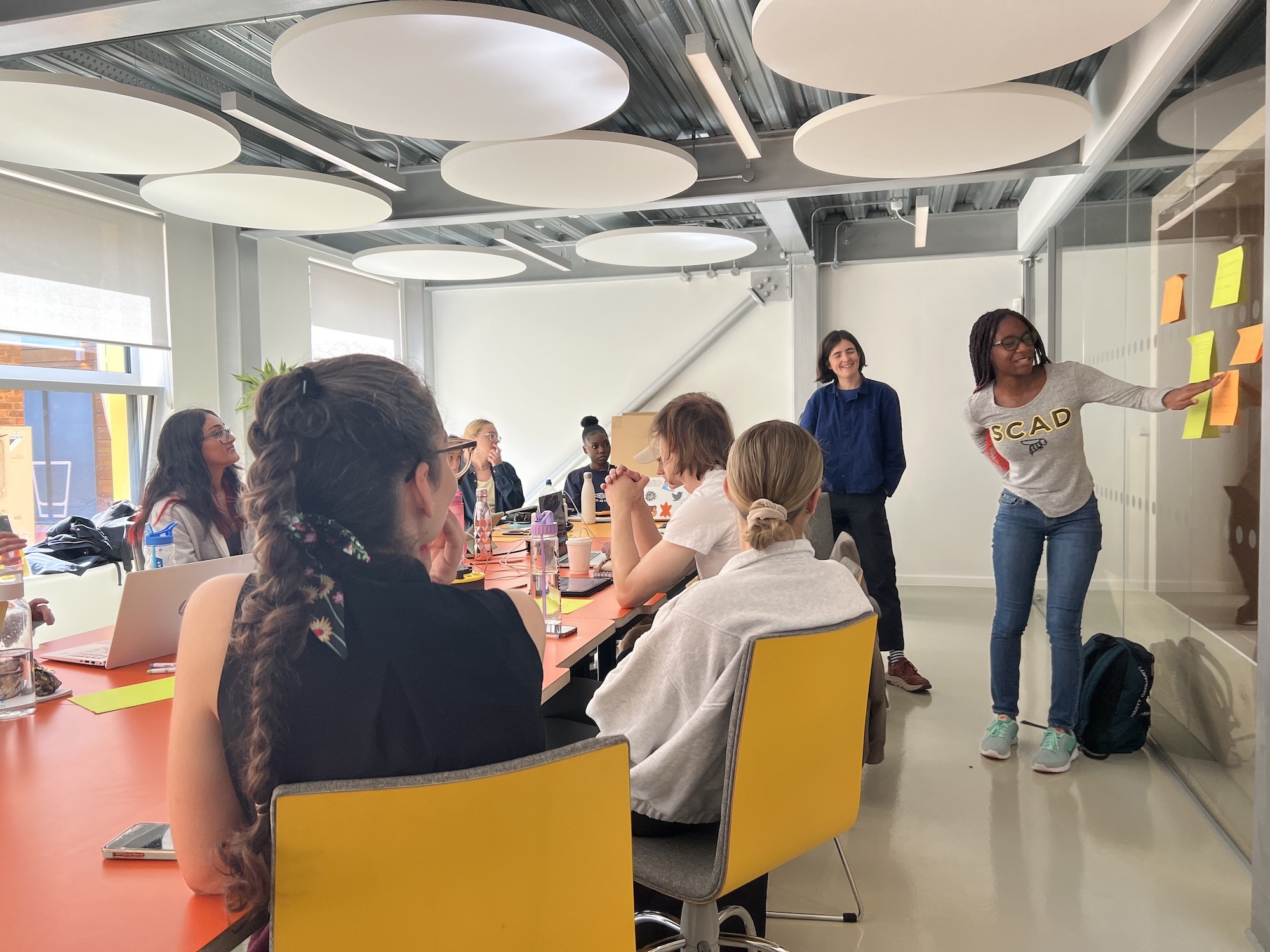 A group of people sitting around a large table in the Innovation Studio. Two people are standing by the table delivering a presentation, with pieces of coloured paper stuck on the wall.