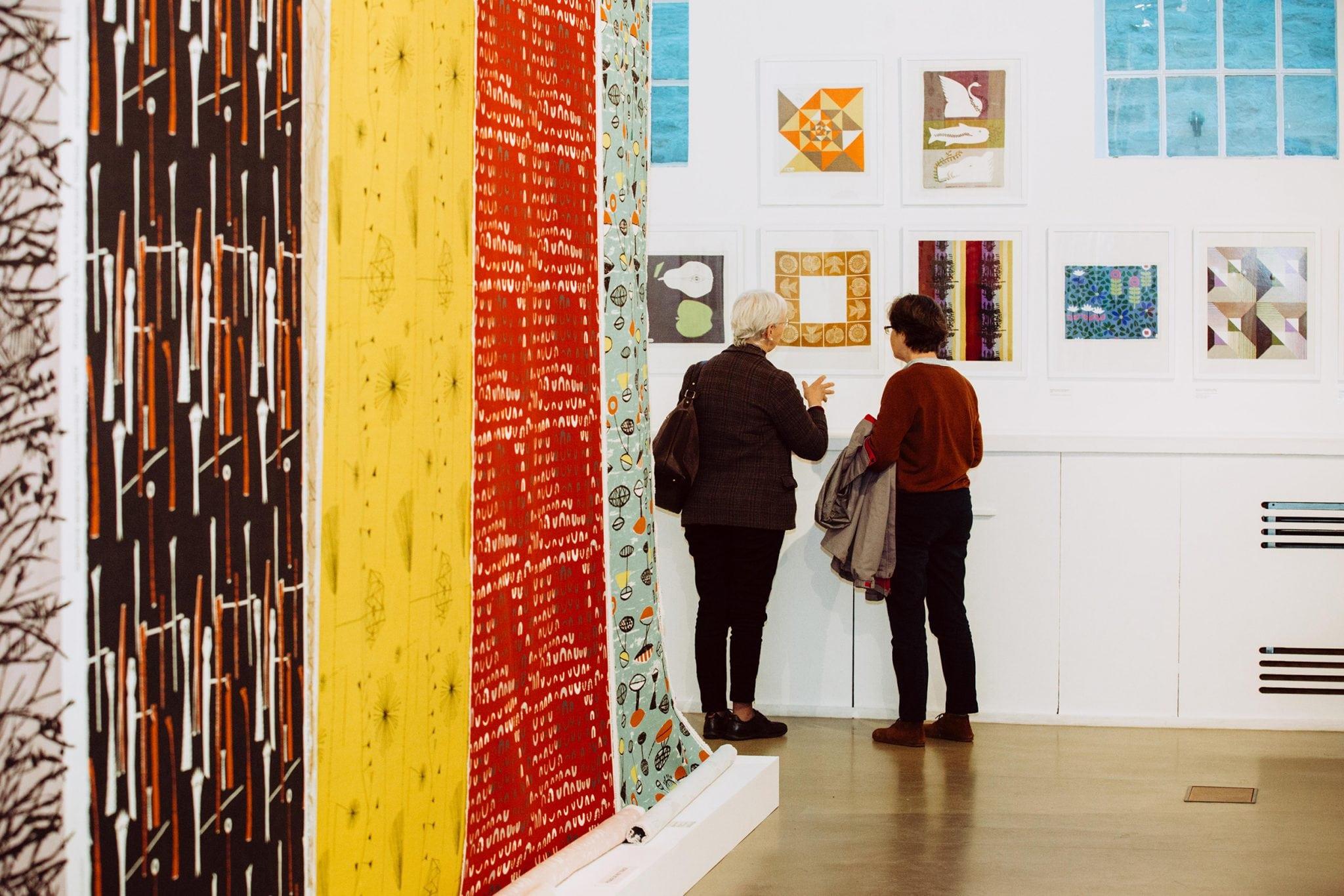 Two people standing in front of a series of artworks hanging on a gallery wall. Long rolls of artwork are hanging on a wall behind them.