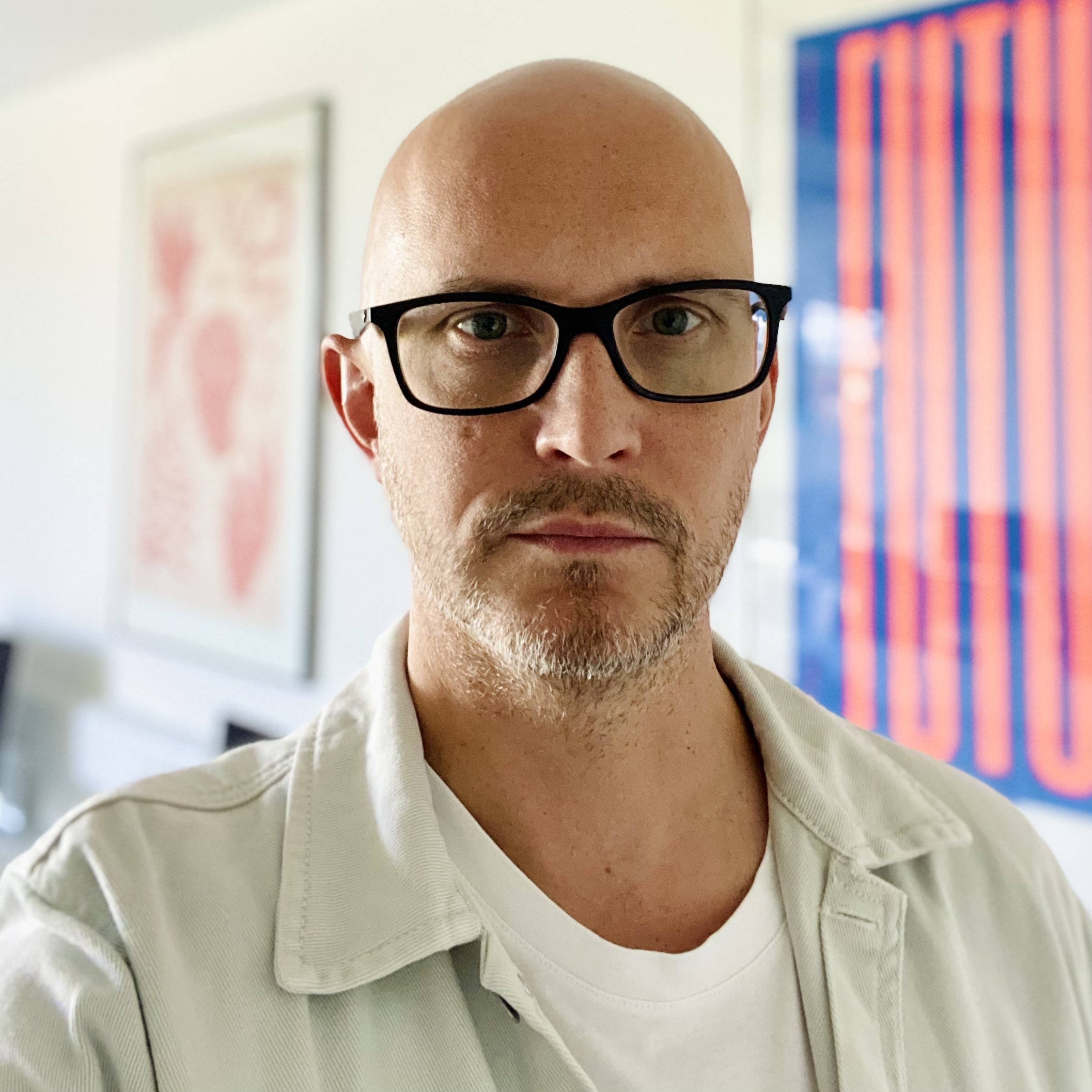 Bald man with black-rimmed glasses and grey beard wearing light green shirt over white t-shirt, colourful artwork visible behind.
