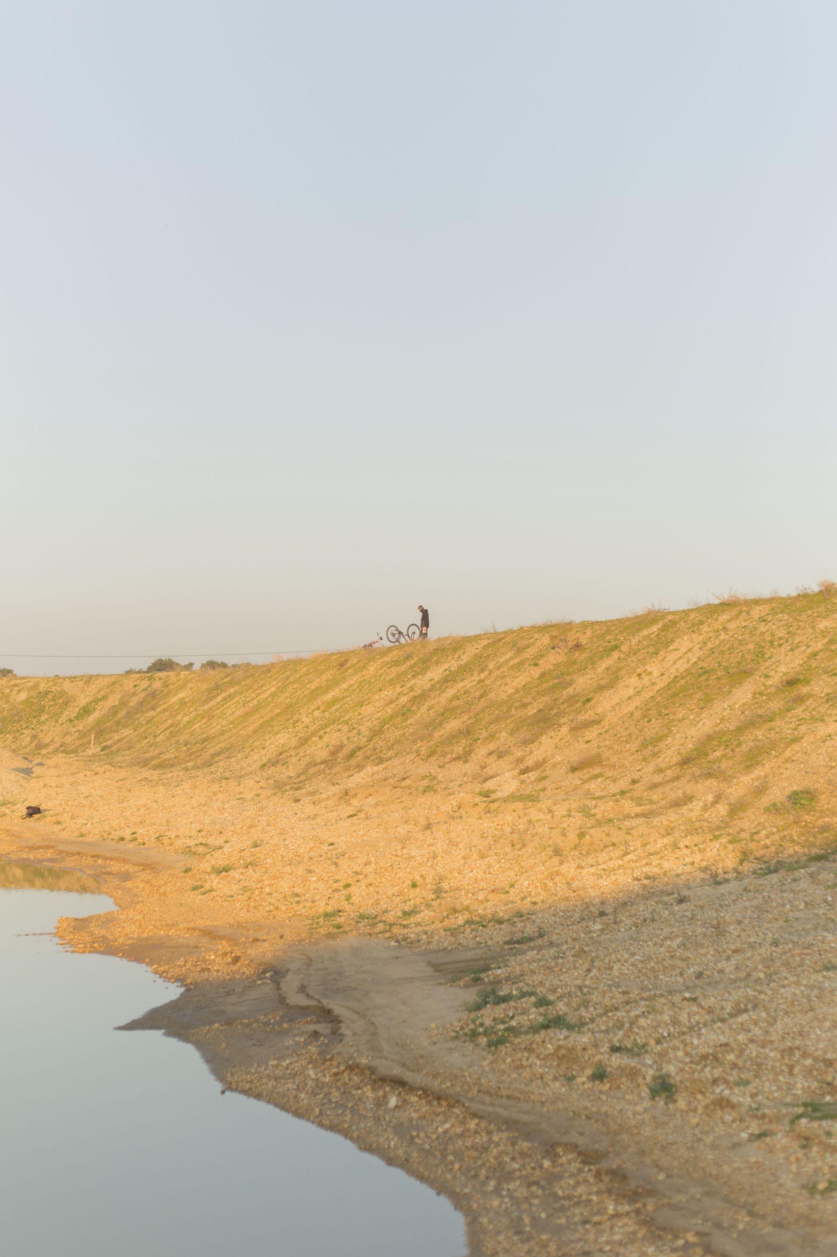 A person with a bicycle standing on a sandy hill in the distance.
