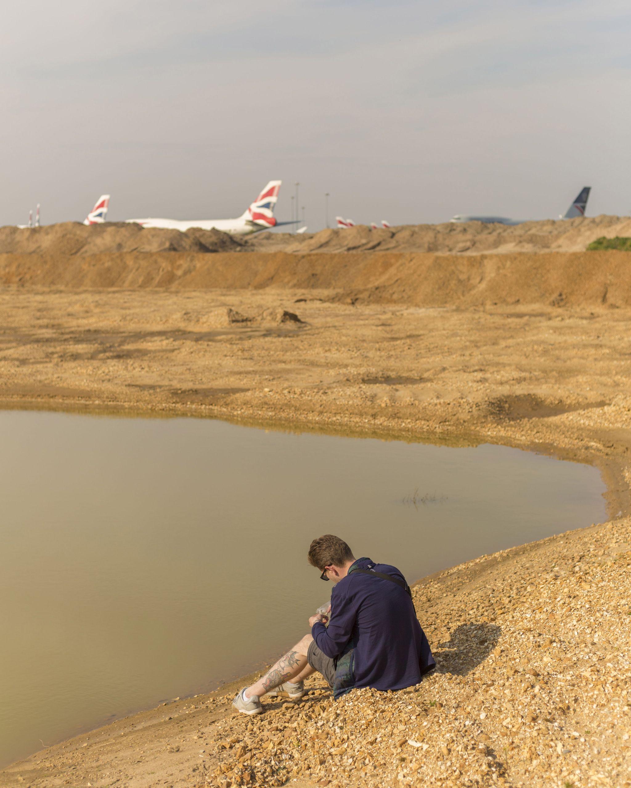 A person sits on a stony beach with sandy hills and aeroplanes in the background