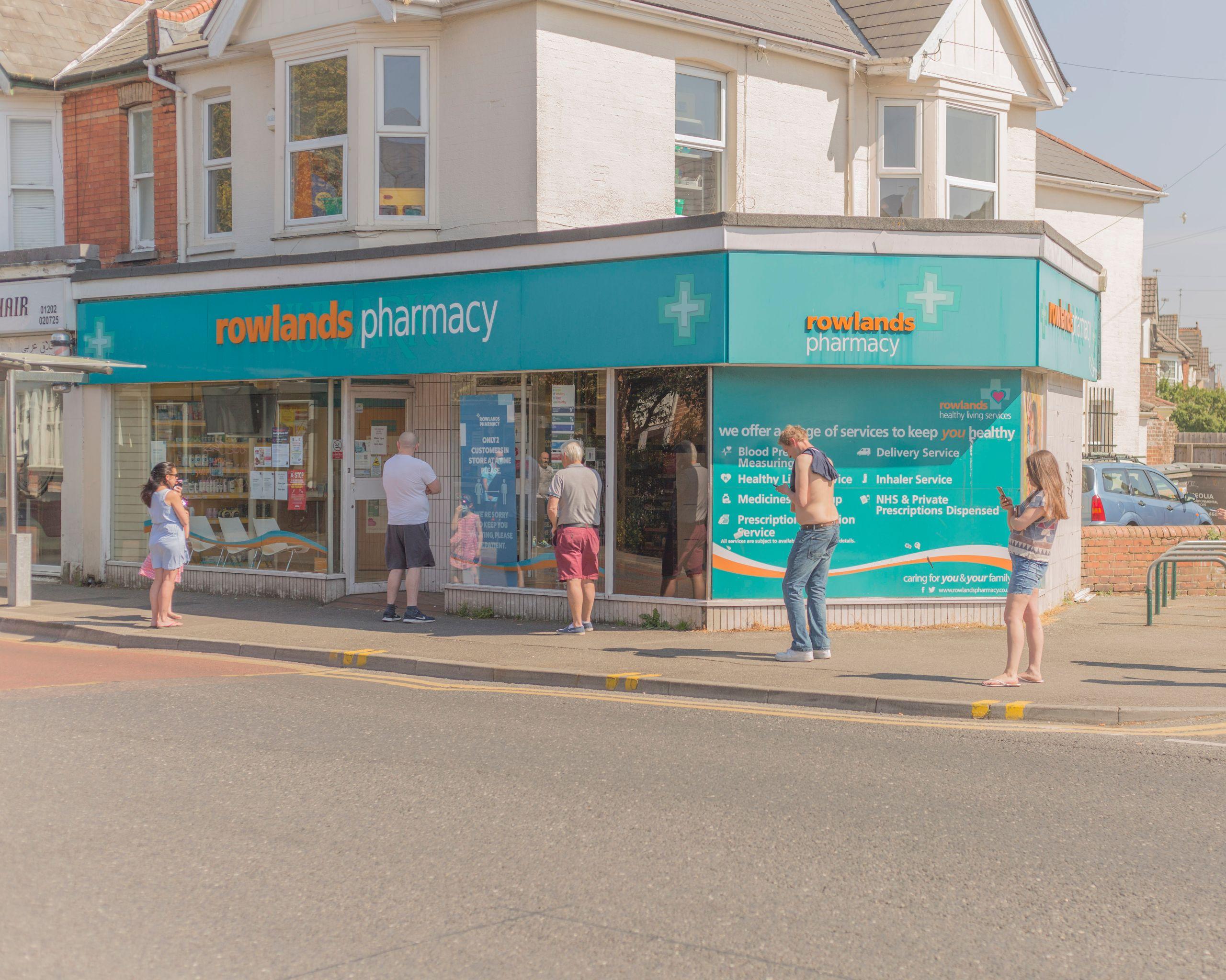 Photo of exterior of Rowlands Pharmacy on a street corner with people outside it.