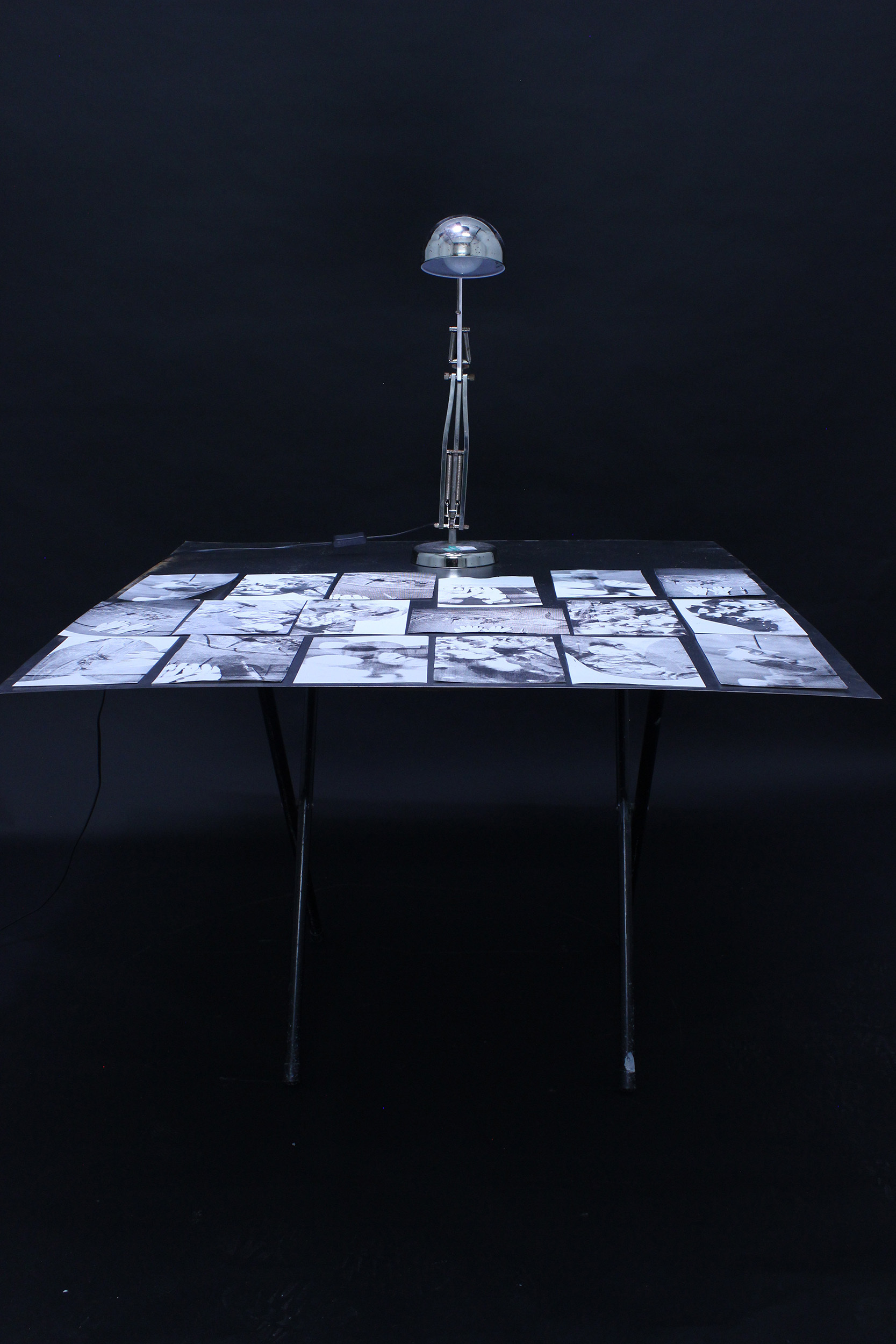 Dark room with illuminated table displaying grid of white photographs, chrome adjustable desk lamp positioned above centre.