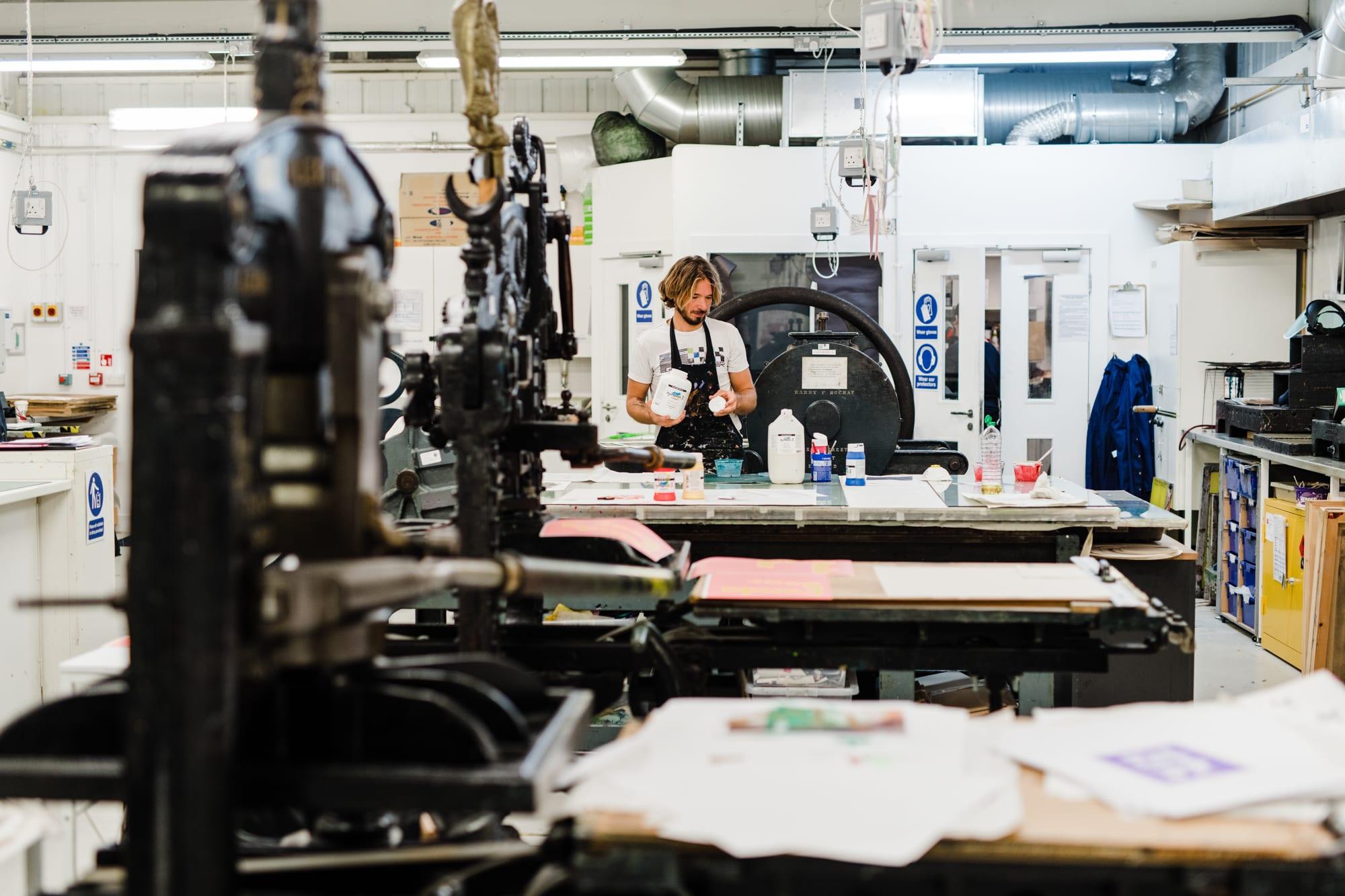 Person stands by a table. Two printing presses are in the foreground.