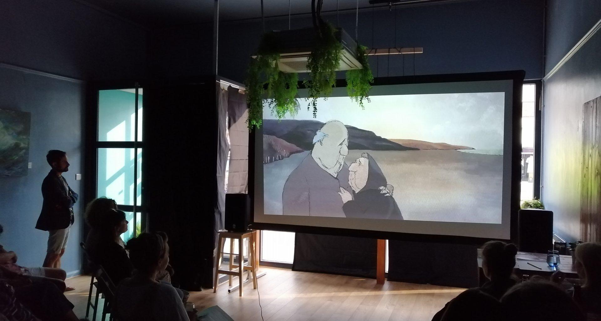 People sit or stand and watch a film in a small cinema space.