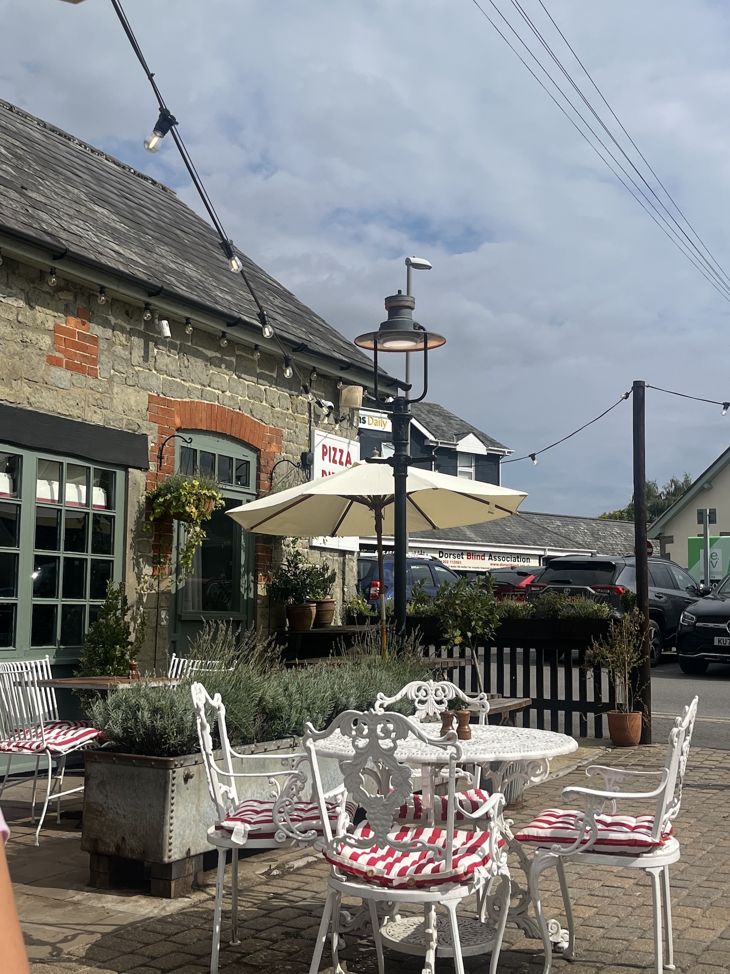 An outdoor café seating area with white metal tables and chairs with red-striped cushions, beside a stone building with hanging lights and an umbrella under a cloudy sky.