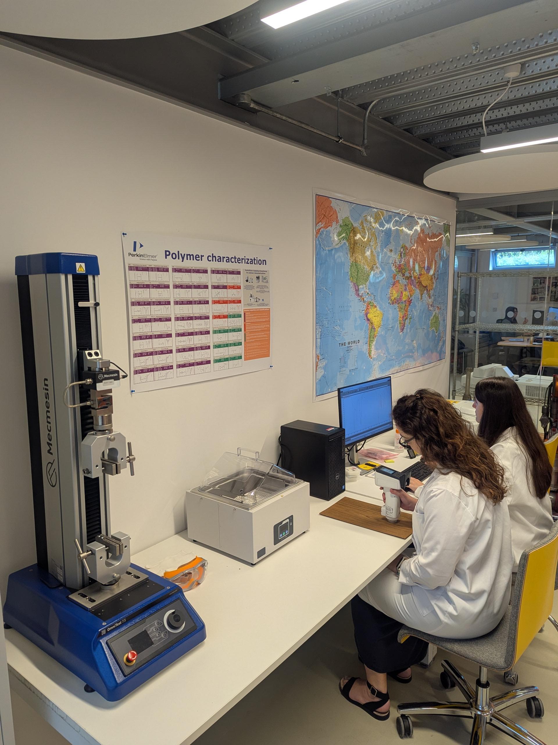 Laboratory with blue testing machine on left, two people in white coats at computer workstation, world map and polymer chart on wall.