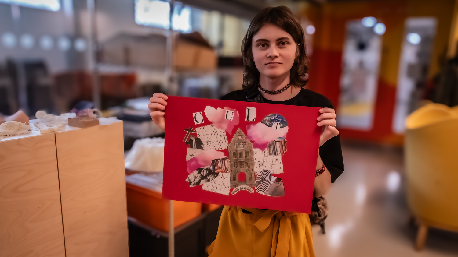 Woman with dark hair holding red fabric artwork featuring architectural imagery in bright indoor space with cardboard boxes.