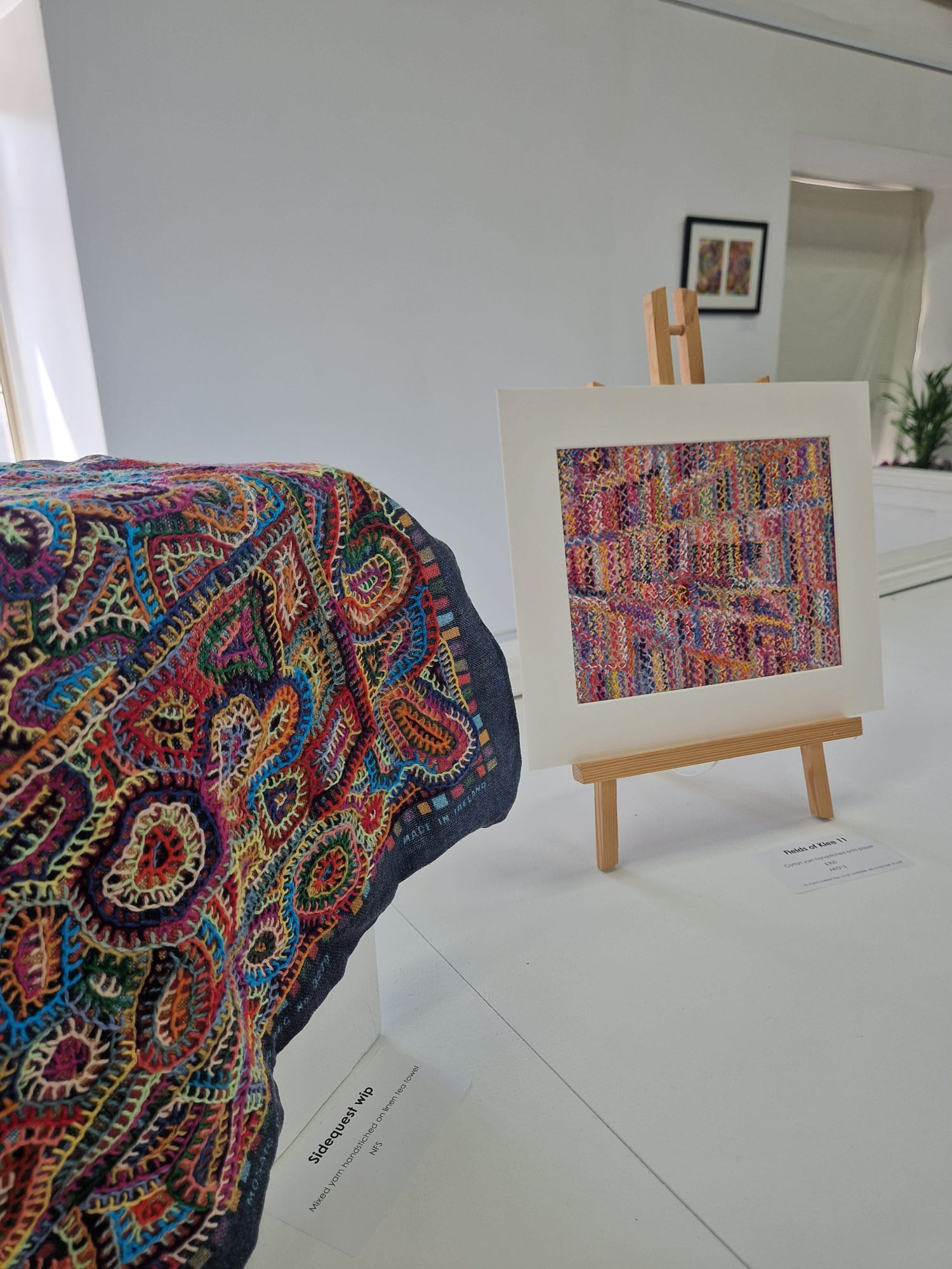 A vibrant textile featuring intricate patterns in various colours rests on a white display surface, while a framed artwork with similar patterns is positioned on an easel nearby. The background consists of a light, neutral-coloured wall.