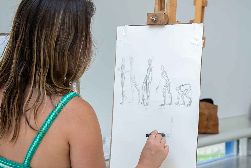 Woman with brown hair wearing green striped top drawing pencil sketches of figures on white paper at wooden easel