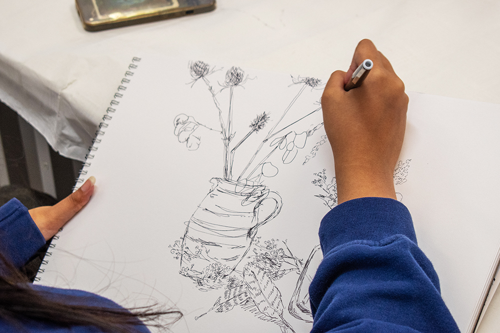 Person in blue sleeves drawing botanical sketch of flowers in vase on spiral notebook with pen
