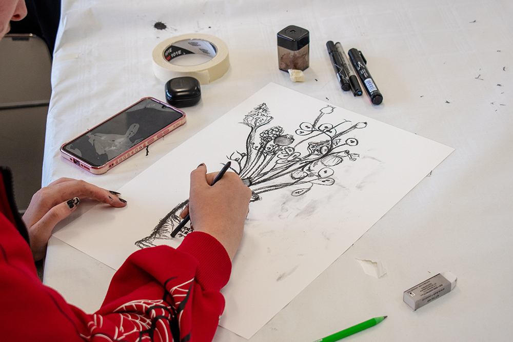 Person in red jumper drawing floral design on white paper with black pen, art supplies and mobile phone on white table