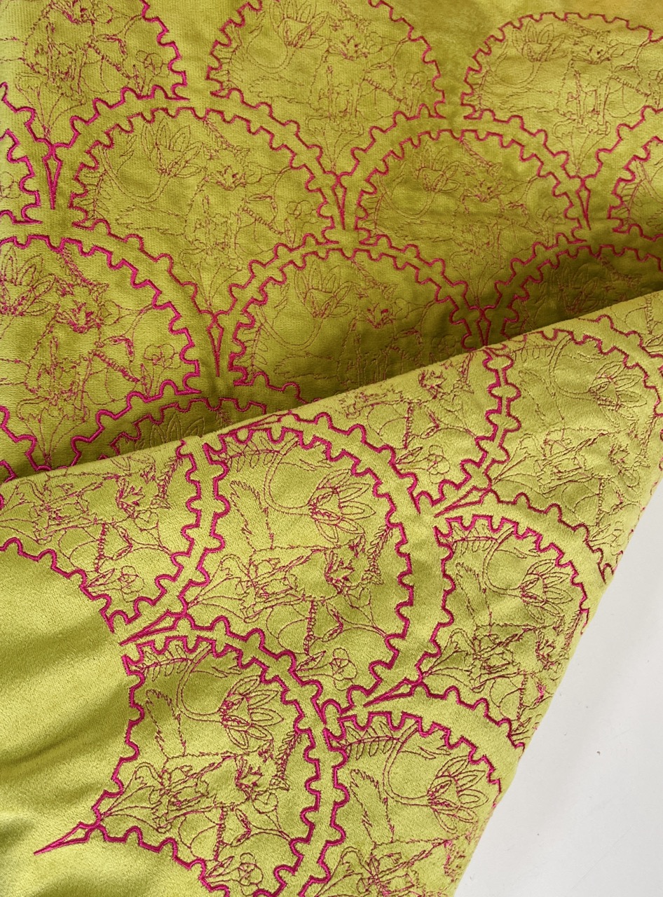 A close-up view of a vibrant green fabric featuring intricate patterns of circular motifs outlined in pink. The fabric has a smooth texture, and portions of it are folded to reveal different sections of the design.
