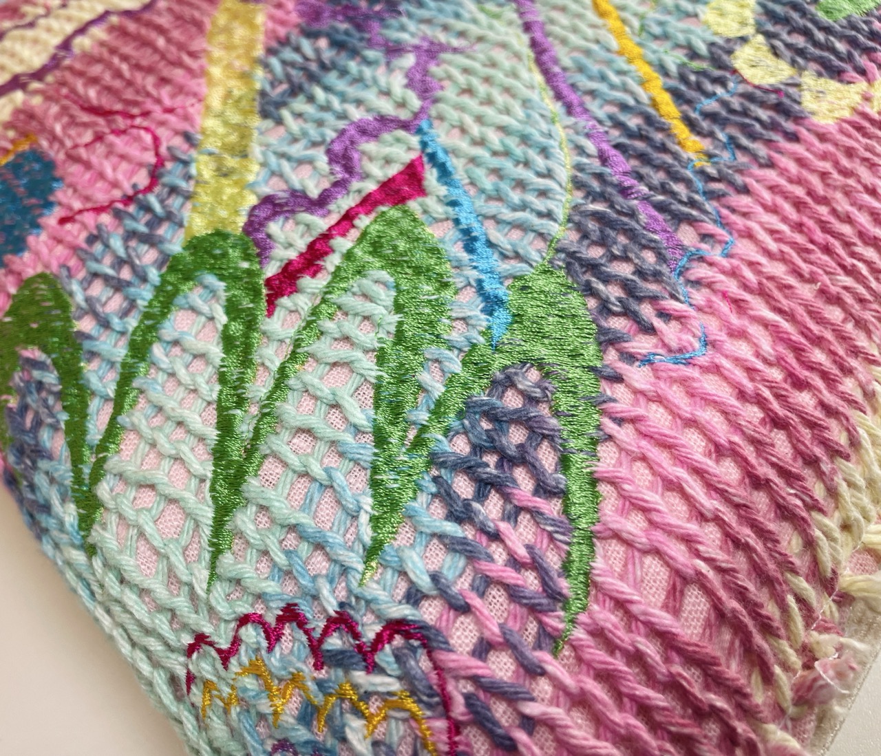 A close-up view of intricately embroidered fabric featuring a blend of pastel colours, including pink, blue, and green. The design includes abstract patterns and varying thread textures, highlighting craftsmanship in textile art.
