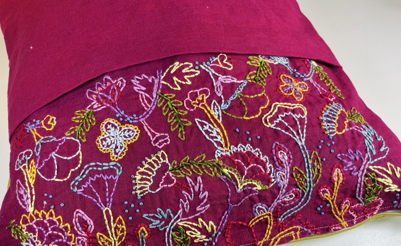 A close-up of a maroon cushion featuring intricate floral embroidery at the bottom. The embroidered design includes various colourful flowers and leaves in shades of yellow, green, and pink against the maroon fabric.